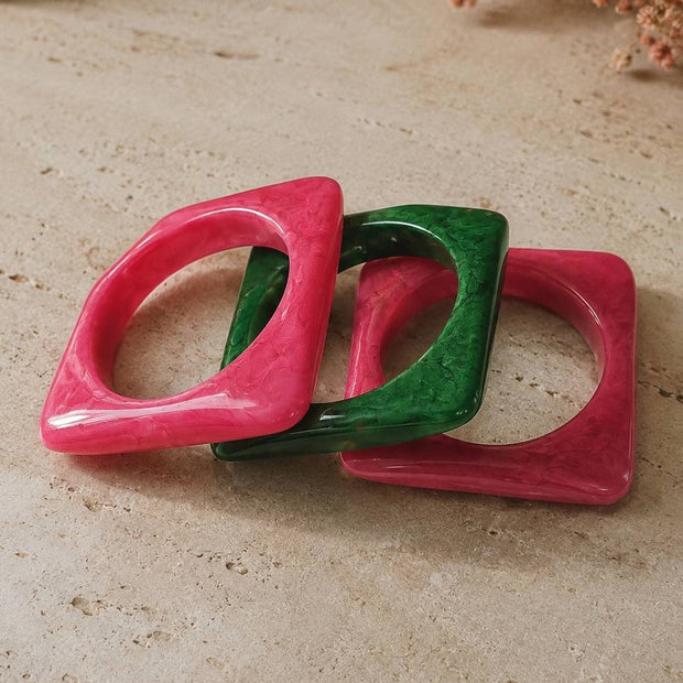 Three square marble-finish resin bangles in hot pink and emerald green arranged overlapping on a stone surface with dried flowers