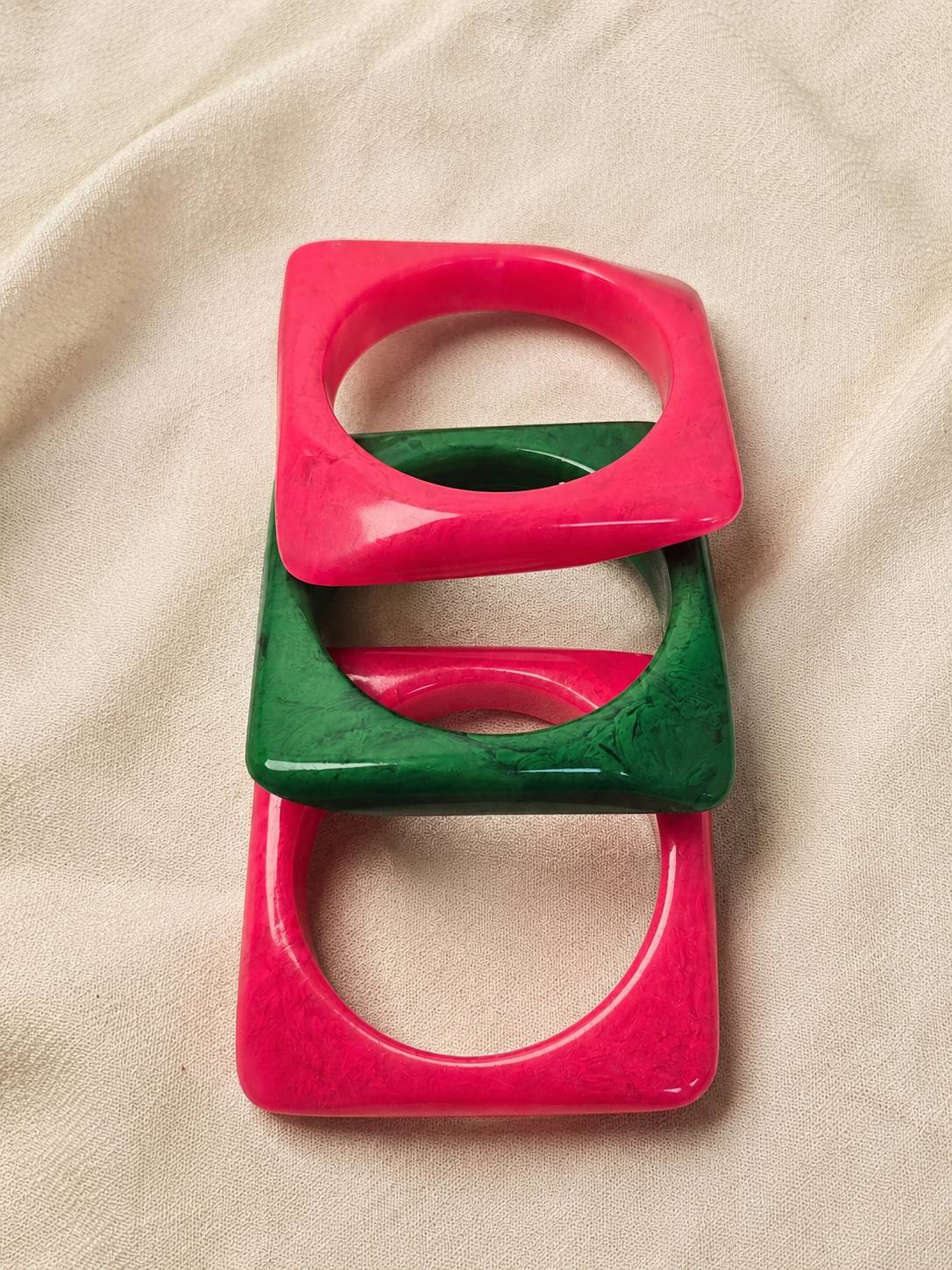 Stack of three square resin bangles in pink and green laid flat on cream linen fabric