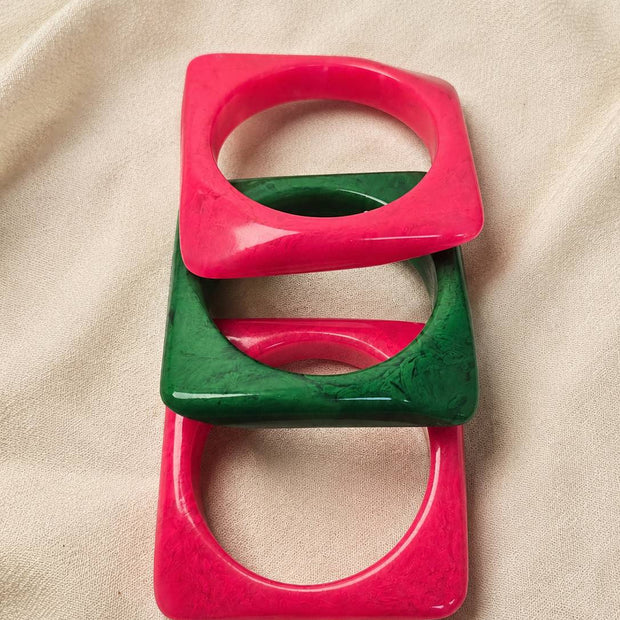 Stack of three square resin bangles in pink and green laid flat on cream linen fabric