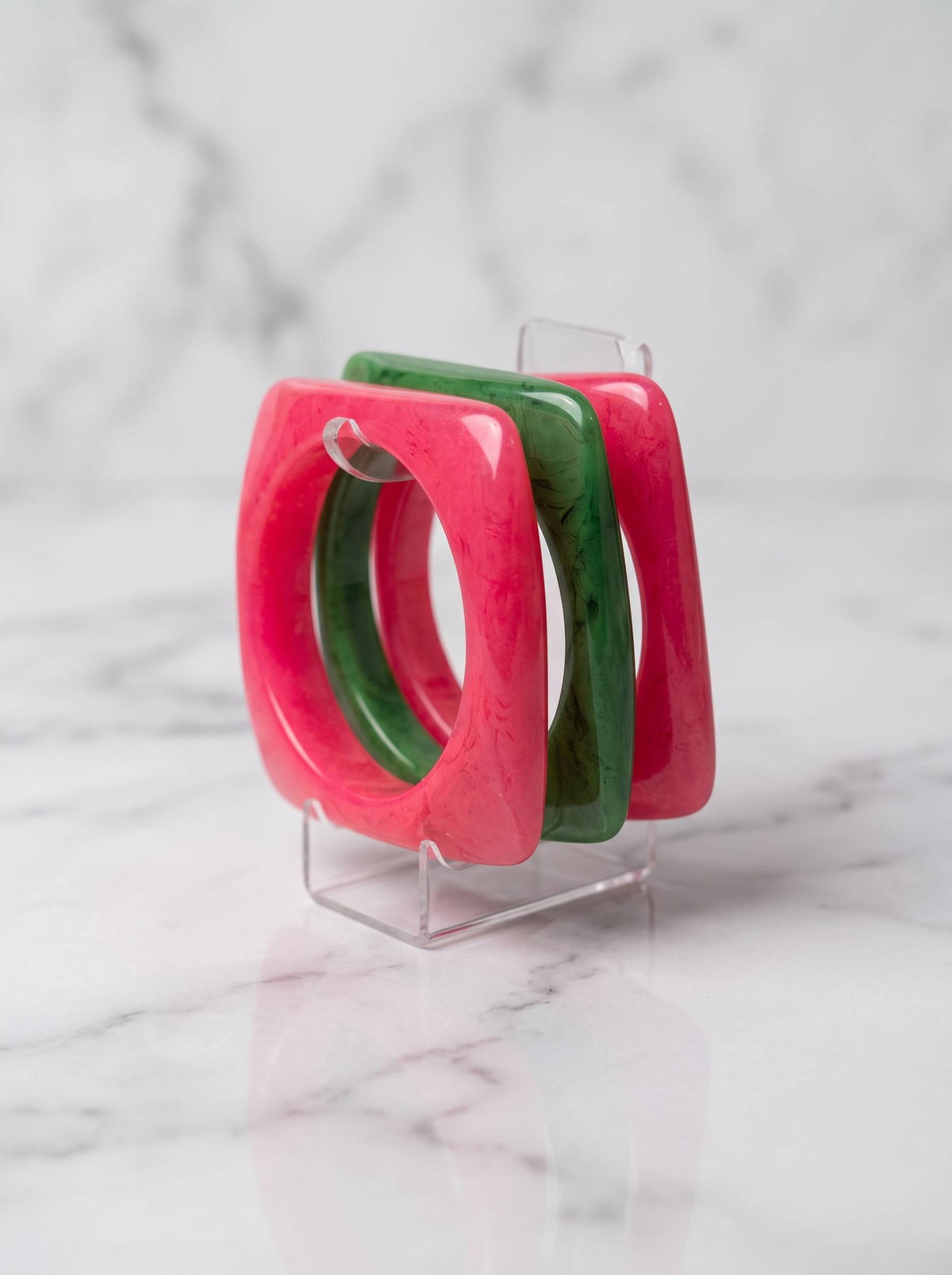 Hot pink and emerald green square resin bangles displayed upright on a clear acrylic stand against a marble background