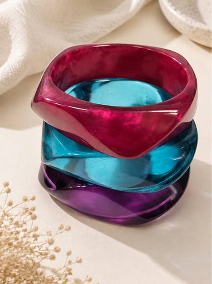 Three jewel-toned acrylic twisted bangles stacked vertically in magenta, teal and purple on a cream surface