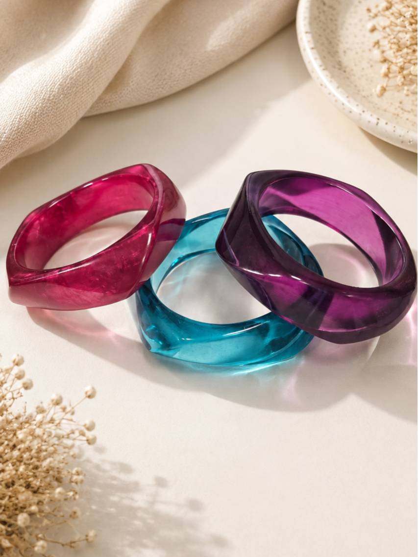 Magenta, teal and purple acrylic twisted bangles laid flat and fanned out on a white surface with dried flowers