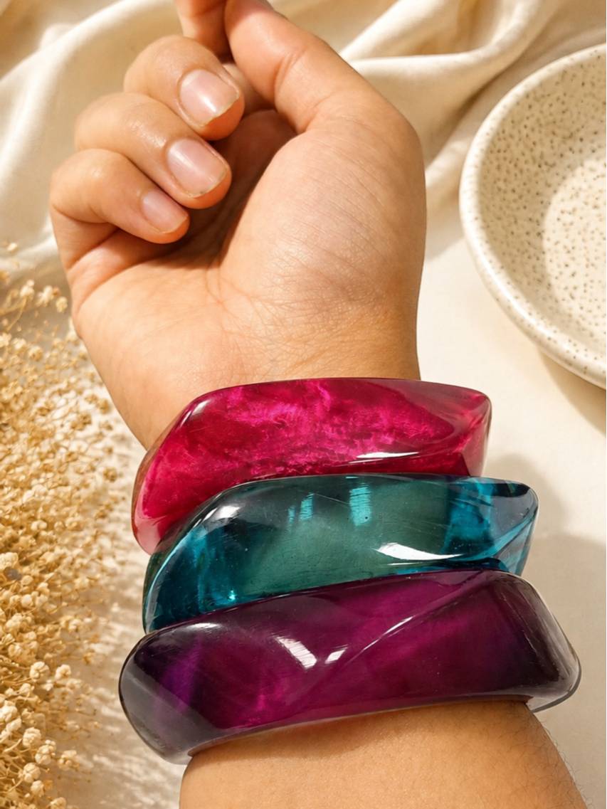 Close-up of three acrylic twisted bangles worn together on a wrist, showing magenta, teal and purple stacked