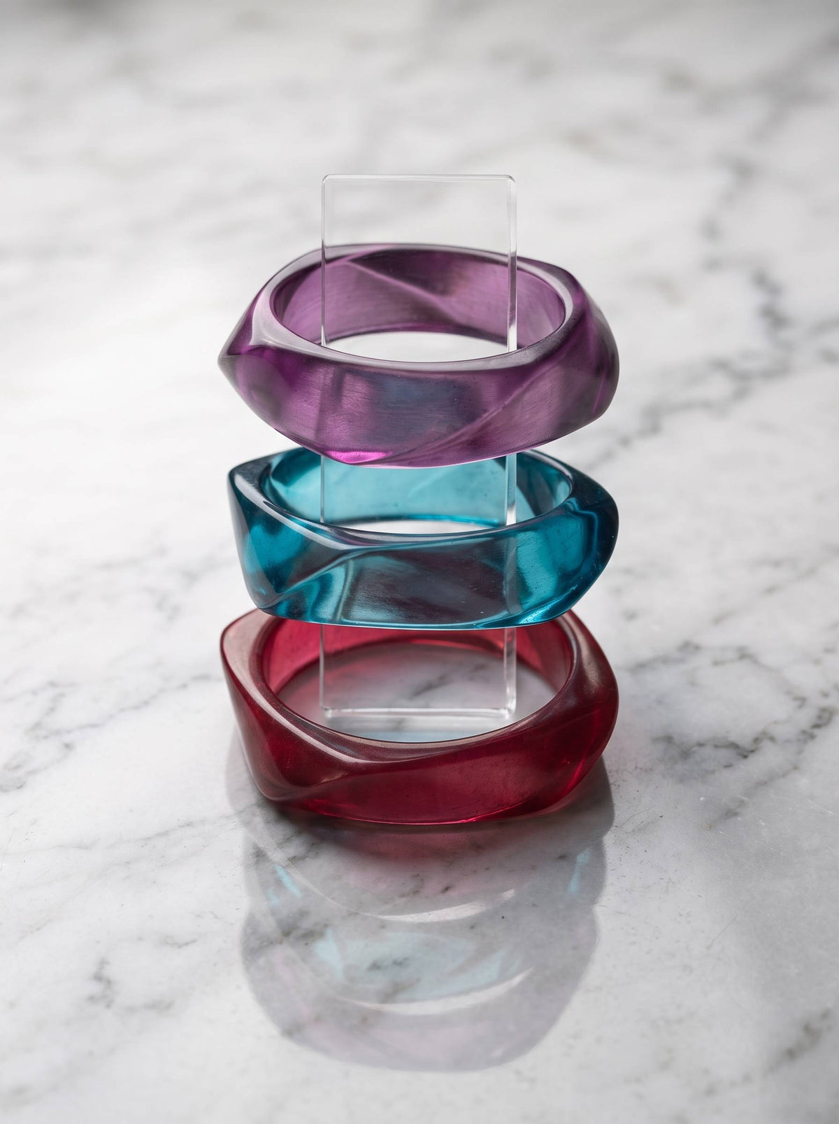 Three acrylic twisted bangles in purple, teal and magenta stacked on a clear display stand on marble surface