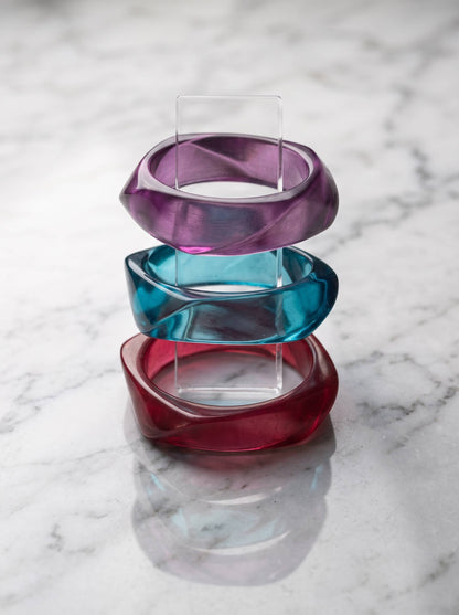 Three acrylic twisted bangles in purple, teal and magenta stacked on a clear display stand on marble surface