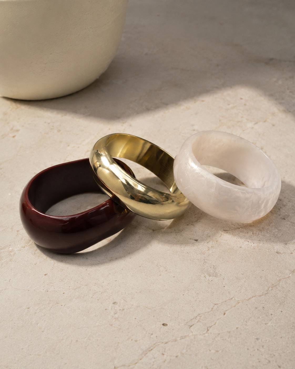 Three chunky dome bangles in burgundy resin, gold brass and white pearl resin resting on a marble surface beside a white vase