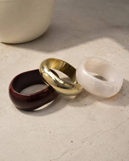 Three chunky dome bangles in burgundy resin, gold brass and white pearl resin resting on a marble surface beside a white vase