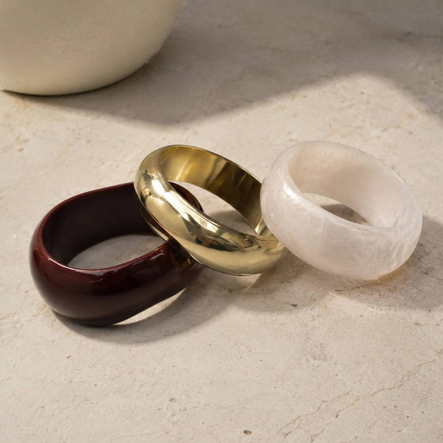 Three chunky dome bangles in burgundy resin, gold brass and white pearl resin resting on a marble surface beside a white vase