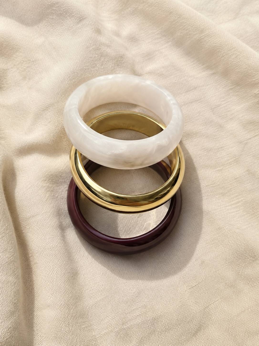 Burgundy resin, gold-finish brass and white pearl resin bangles stacked flat on a cream linen fabric background