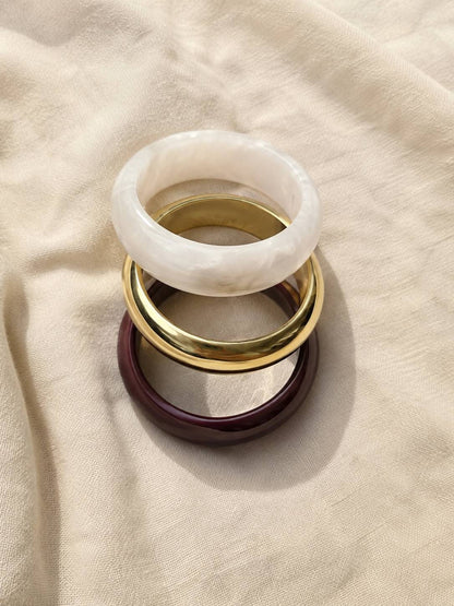Burgundy resin, gold-finish brass and white pearl resin bangles stacked flat on a cream linen fabric background