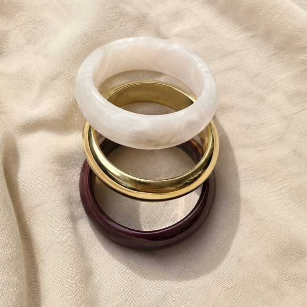 Burgundy resin, gold-finish brass and white pearl resin bangles stacked flat on a cream linen fabric background