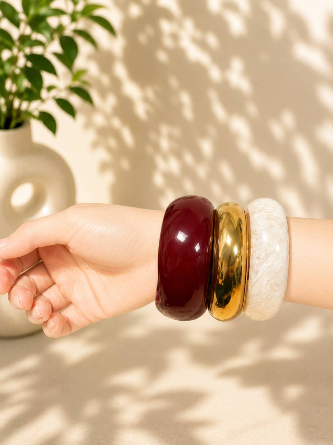 Three chunky stacked bangles in burgundy, gold and white worn on a wrist with a potted plant in the background