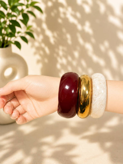 Three chunky stacked bangles in burgundy, gold and white worn on a wrist with a potted plant in the background
