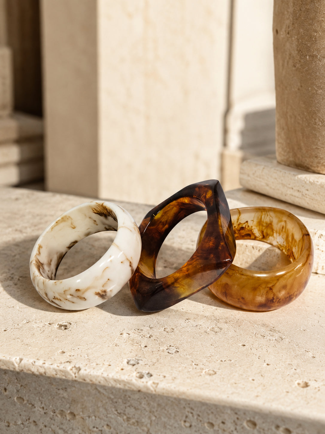 Three resin marble bangles in ivory, dark tortoiseshell, and amber resting on a travertine stone surface