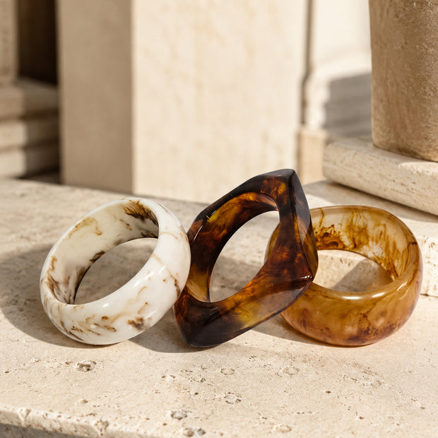 Three resin marble bangles in ivory, dark tortoiseshell, and amber resting on a travertine stone surface