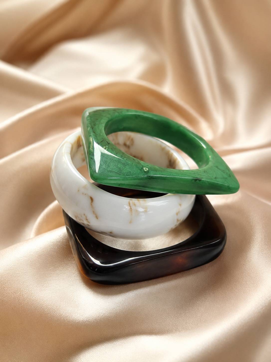 Three marble resin bangles stacked on gold satin — green geometric, ivory marbled, and dark tortoiseshell square