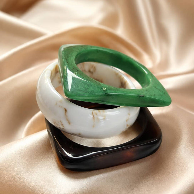Three marble resin bangles stacked on gold satin — green geometric, ivory marbled, and dark tortoiseshell square