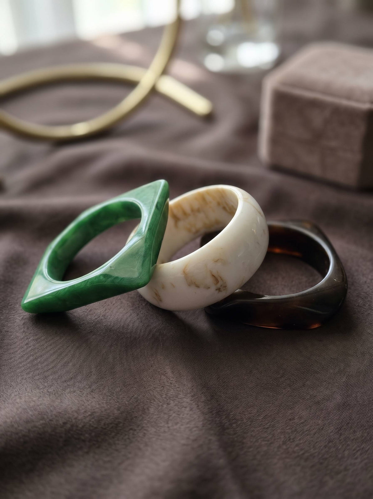 Green diamond-shaped, ivory round, and tortoiseshell square resin bangles laid flat on grey fabric