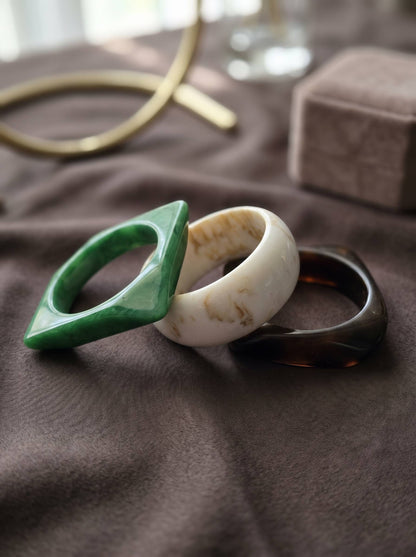 Green diamond-shaped, ivory round, and tortoiseshell square resin bangles laid flat on grey fabric