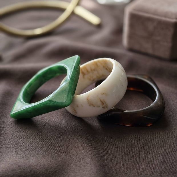 Green diamond-shaped, ivory round, and tortoiseshell square resin bangles laid flat on grey fabric