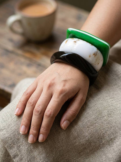 Three marble resin bangles worn stacked on a woman's wrist resting on a linen surface near a coffee cup