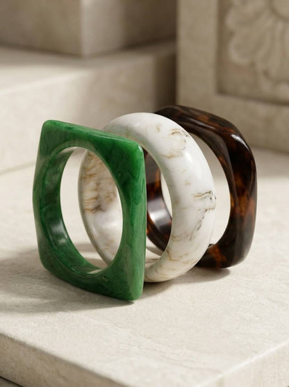 Green, white marbled, and tortoiseshell square resin bangles standing upright against a stone background