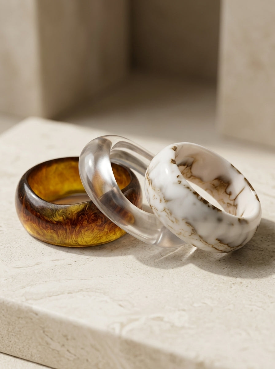 Three chunky resin bangles in amber tortoise, clear crystal, and white marble arranged on a stone surface