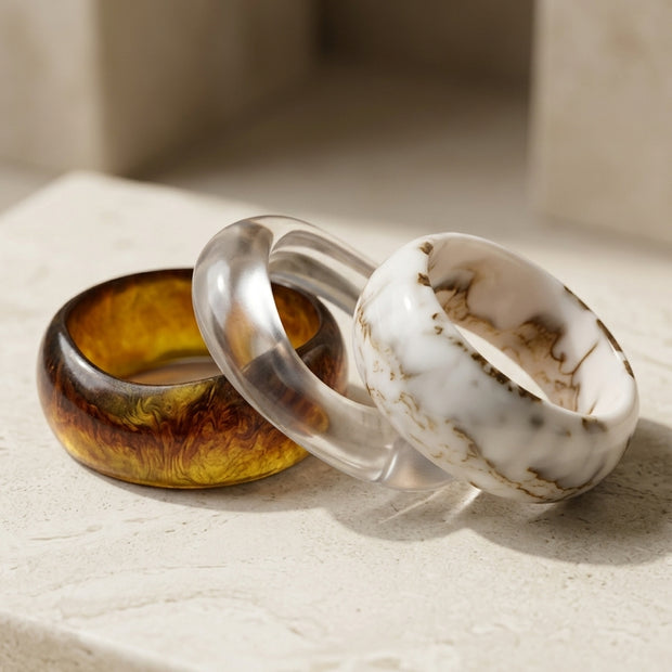 Three chunky resin bangles in amber tortoise, clear crystal, and white marble arranged on a stone surface