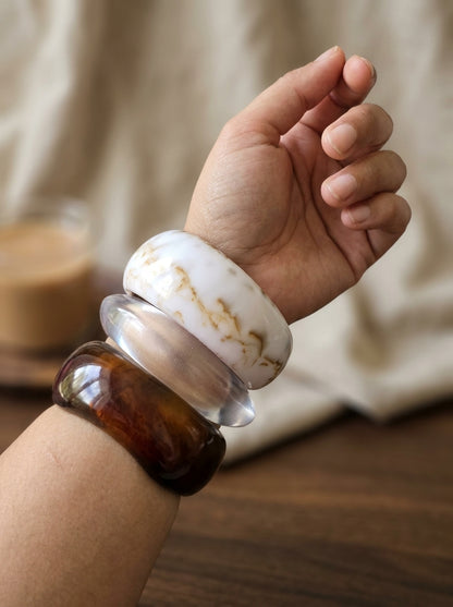 Three resin bangles worn together on a wrist — white marble, clear, and amber tortoiseshell stacked