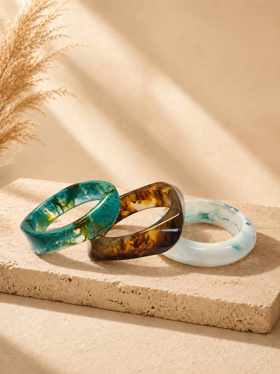 Three resin marble bangles in teal, amber tortoise and white blue laid flat on a sandy stone surface with dried pampas grass