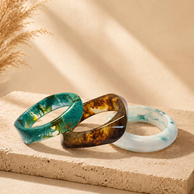 Three resin marble bangles in teal, amber tortoise and white blue laid flat on a sandy stone surface with dried pampas grass