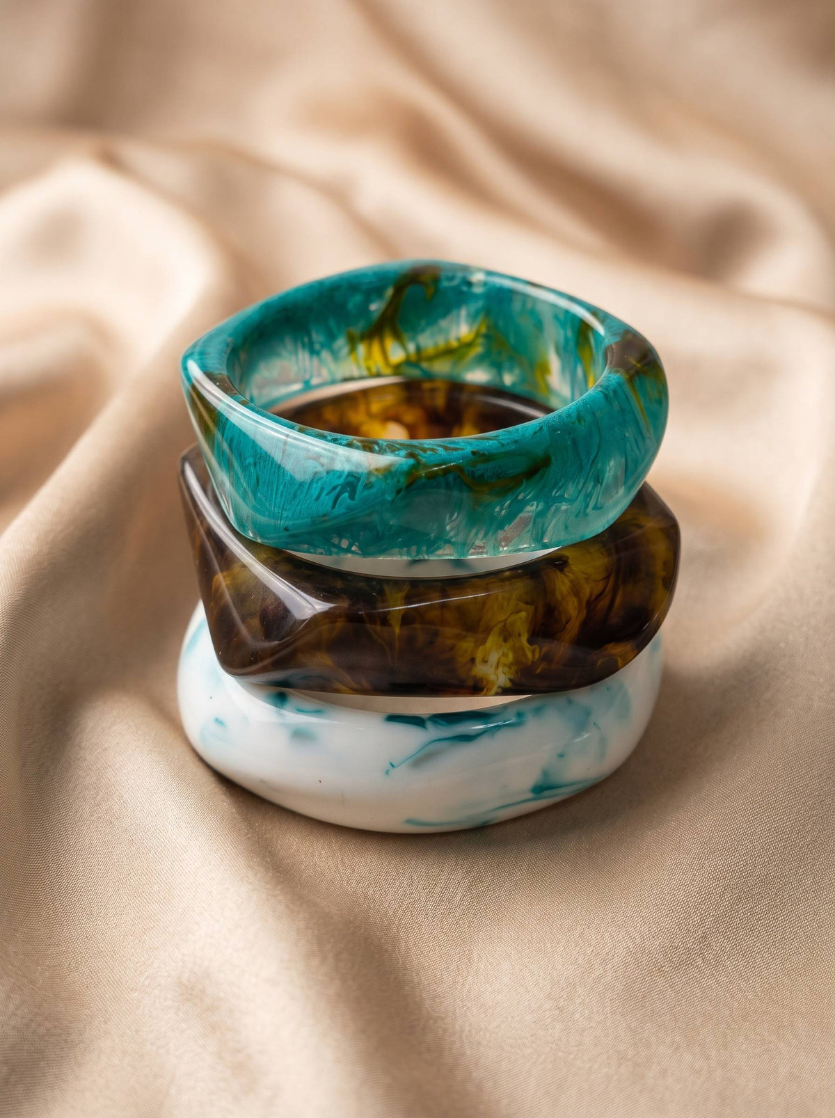 Stack of three resin marble bangles in teal, amber and white piled on cream satin fabric