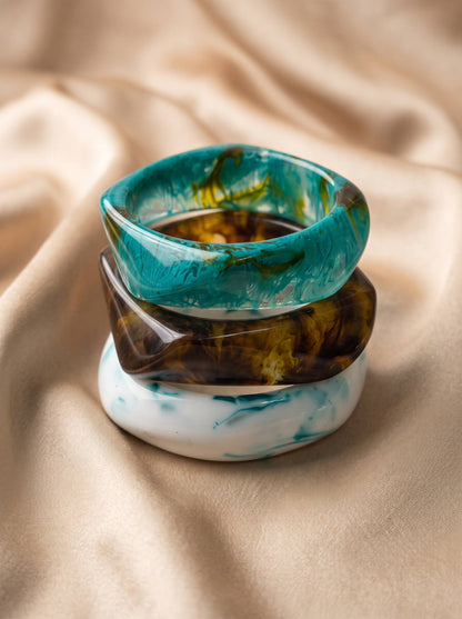 Stack of three resin marble bangles in teal, amber and white piled on cream satin fabric