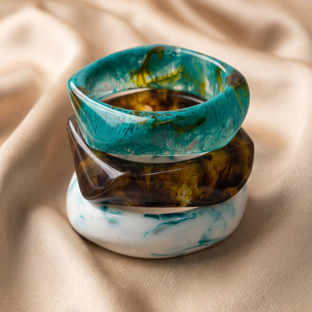 Stack of three resin marble bangles in teal, amber and white piled on cream satin fabric