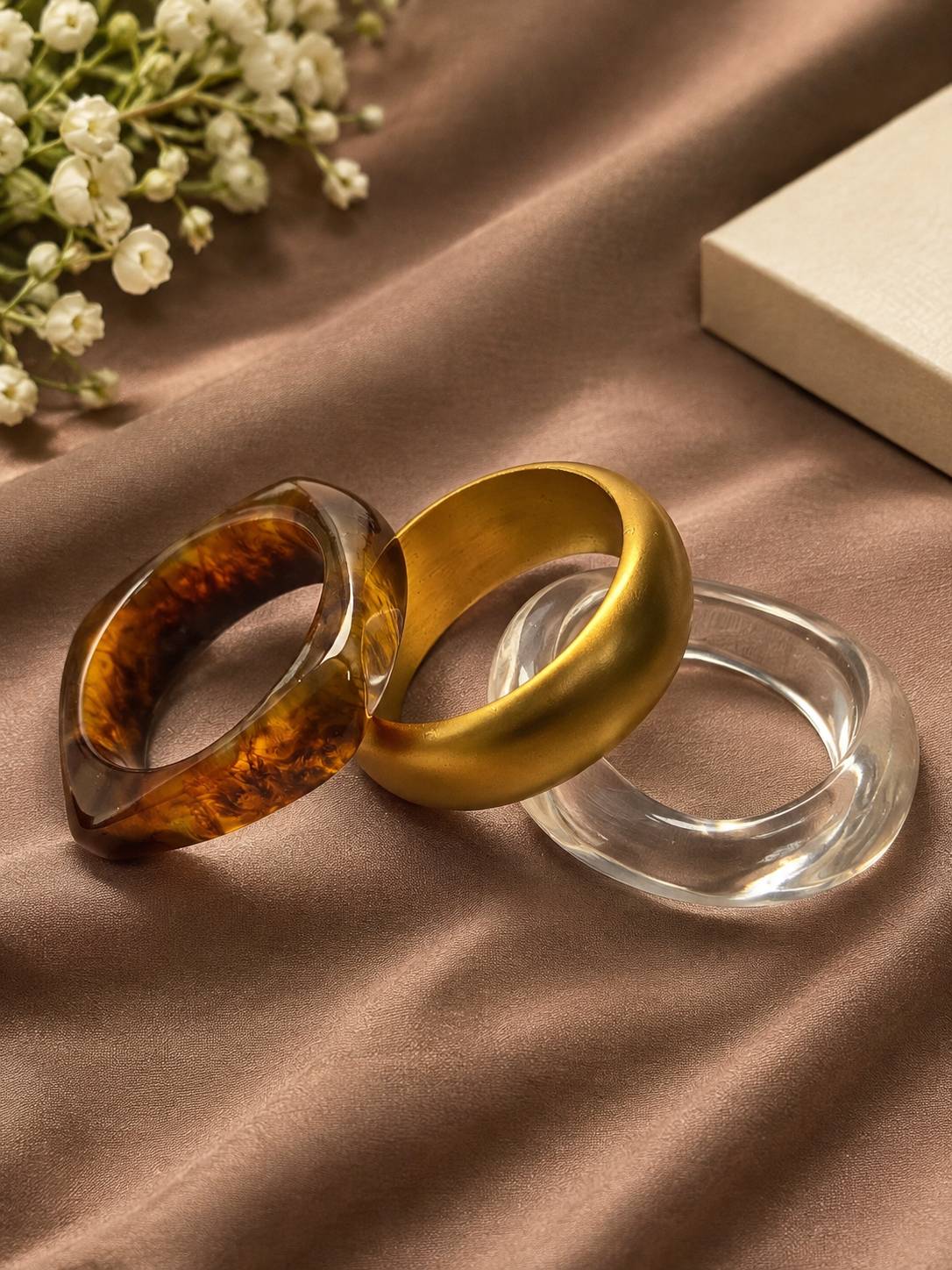 Three bangles laid flat on satin — tortoiseshell resin, matte gold acrylic, and clear acrylic with baby's breath flowers nearby