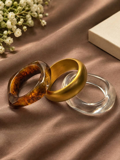 Three bangles laid flat on satin — tortoiseshell resin, matte gold acrylic, and clear acrylic with baby's breath flowers nearby
