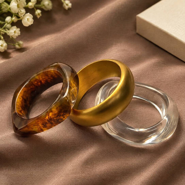 Three bangles laid flat on satin — tortoiseshell resin, matte gold acrylic, and clear acrylic with baby's breath flowers nearby