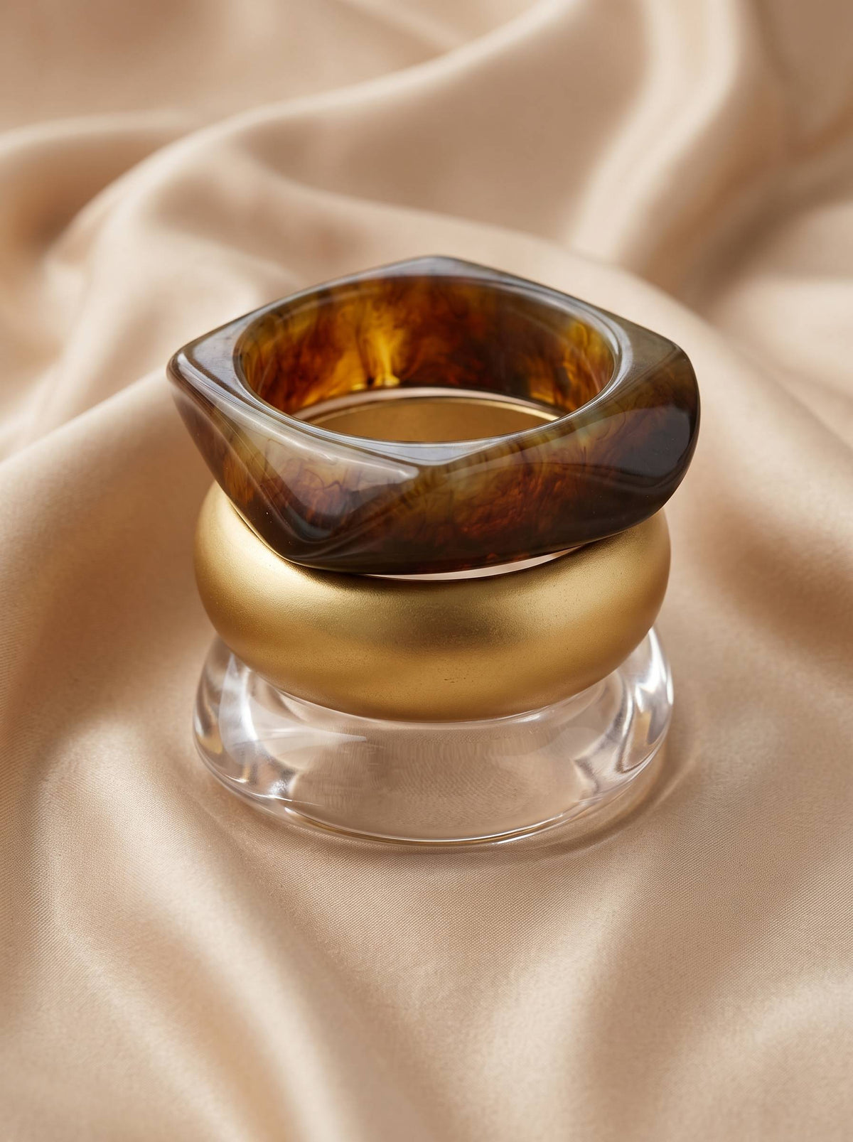 Three bangles stacked upright on cream satin, showing tortoiseshell resin on top, matte gold in middle, clear acrylic at base