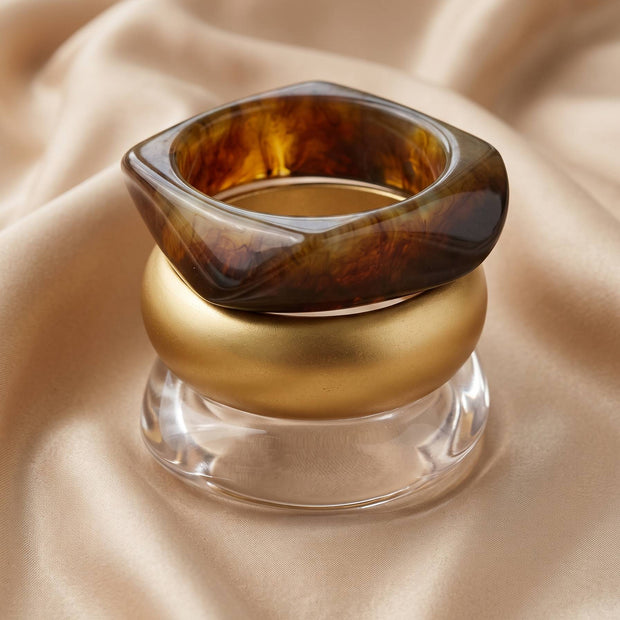 Three bangles stacked upright on cream satin, showing tortoiseshell resin on top, matte gold in middle, clear acrylic at base