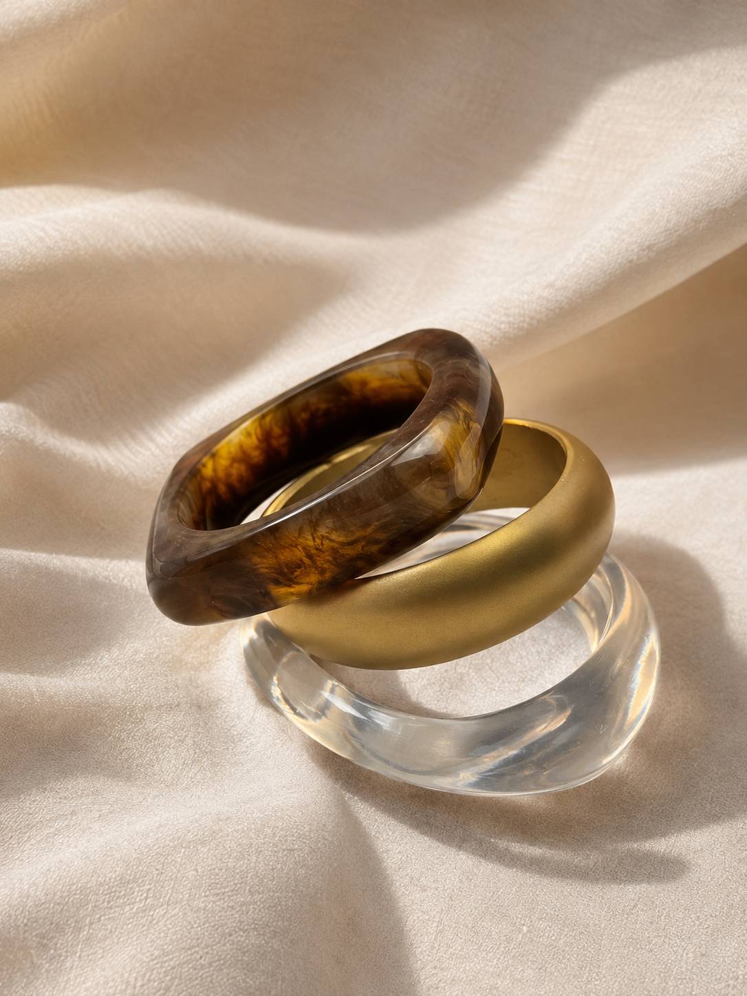 Tortoiseshell resin bangle and matte gold bangle leaning against clear acrylic bangle on cream satin in natural light