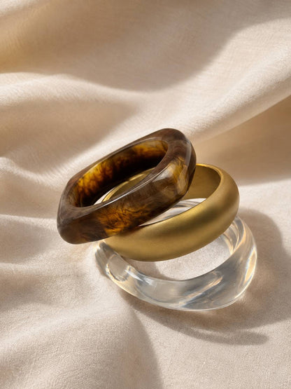 Tortoiseshell resin bangle and matte gold bangle leaning against clear acrylic bangle on cream satin in natural light
