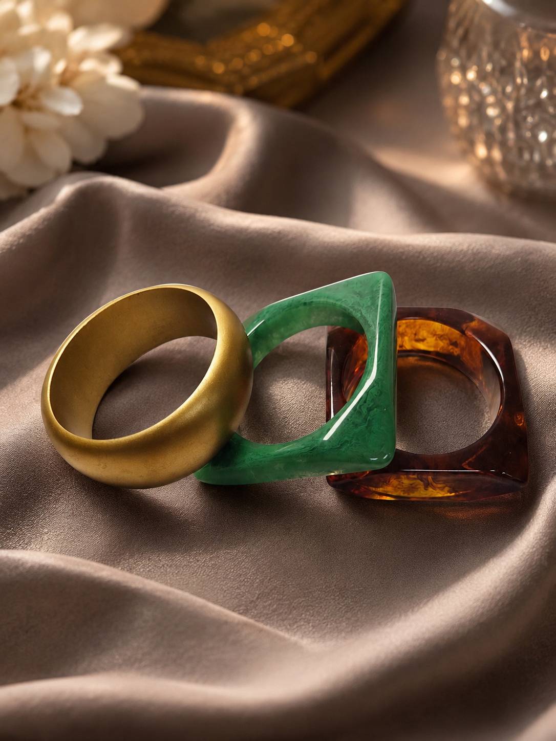 Three stacked bangles laid flat on satin — gold-tone, green marbled resin, and amber tortoiseshell resin