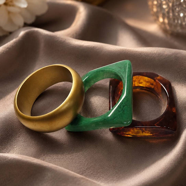Three stacked bangles laid flat on satin — gold-tone, green marbled resin, and amber tortoiseshell resin