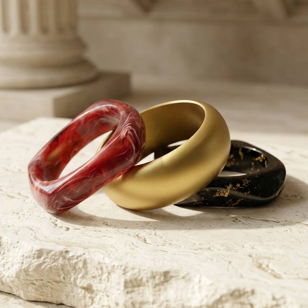 Three resin bangles in red marble, matte gold, and black with gold foil resting on a white stone surface