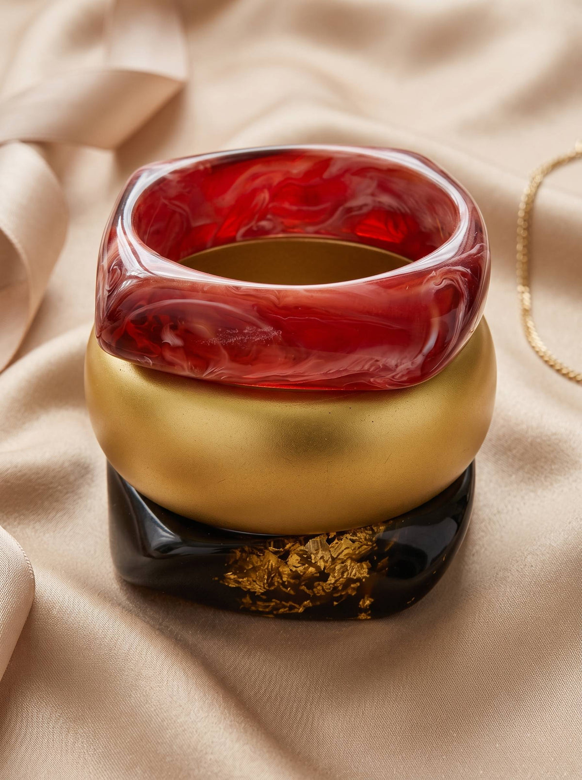 Red marble resin bangle stacked on matte gold and black gold-foil bangles on a satin fabric background