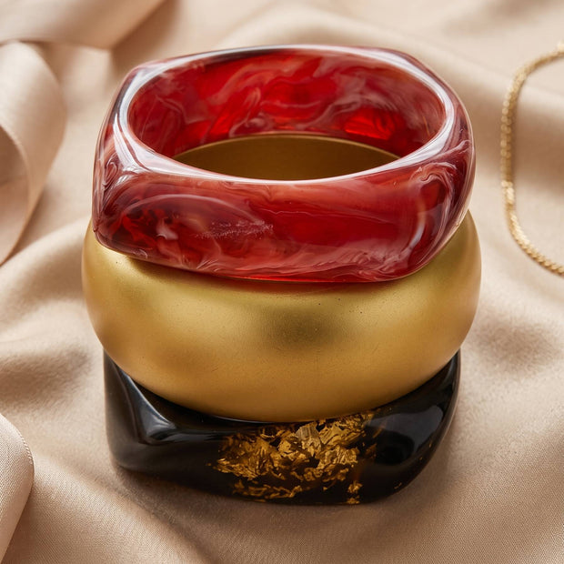 Red marble resin bangle stacked on matte gold and black gold-foil bangles on a satin fabric background