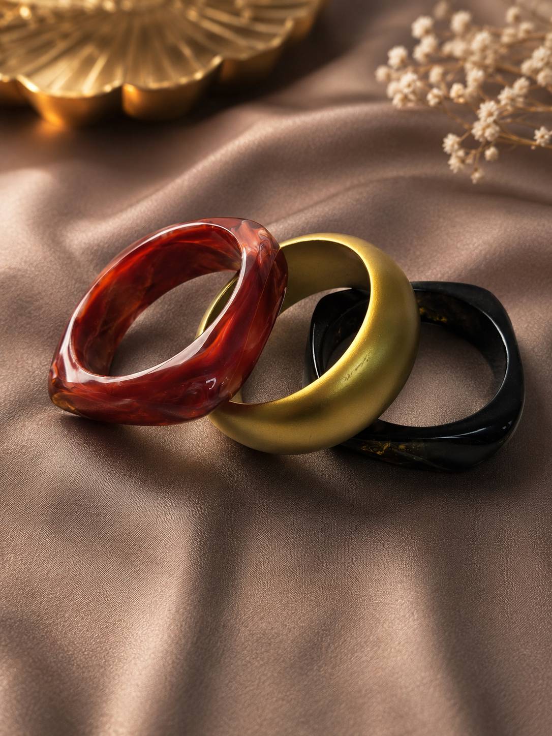 Three resin bangles in red marble, matte gold, and black arranged flat on brown satin fabric with dried flowers