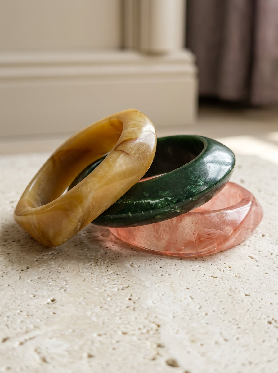 Three marble resin bangles in amber, green, and rose stacked on a stone surface in natural light