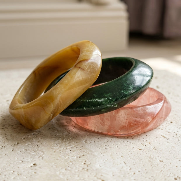 Three marble resin bangles in amber, green, and rose stacked on a stone surface in natural light