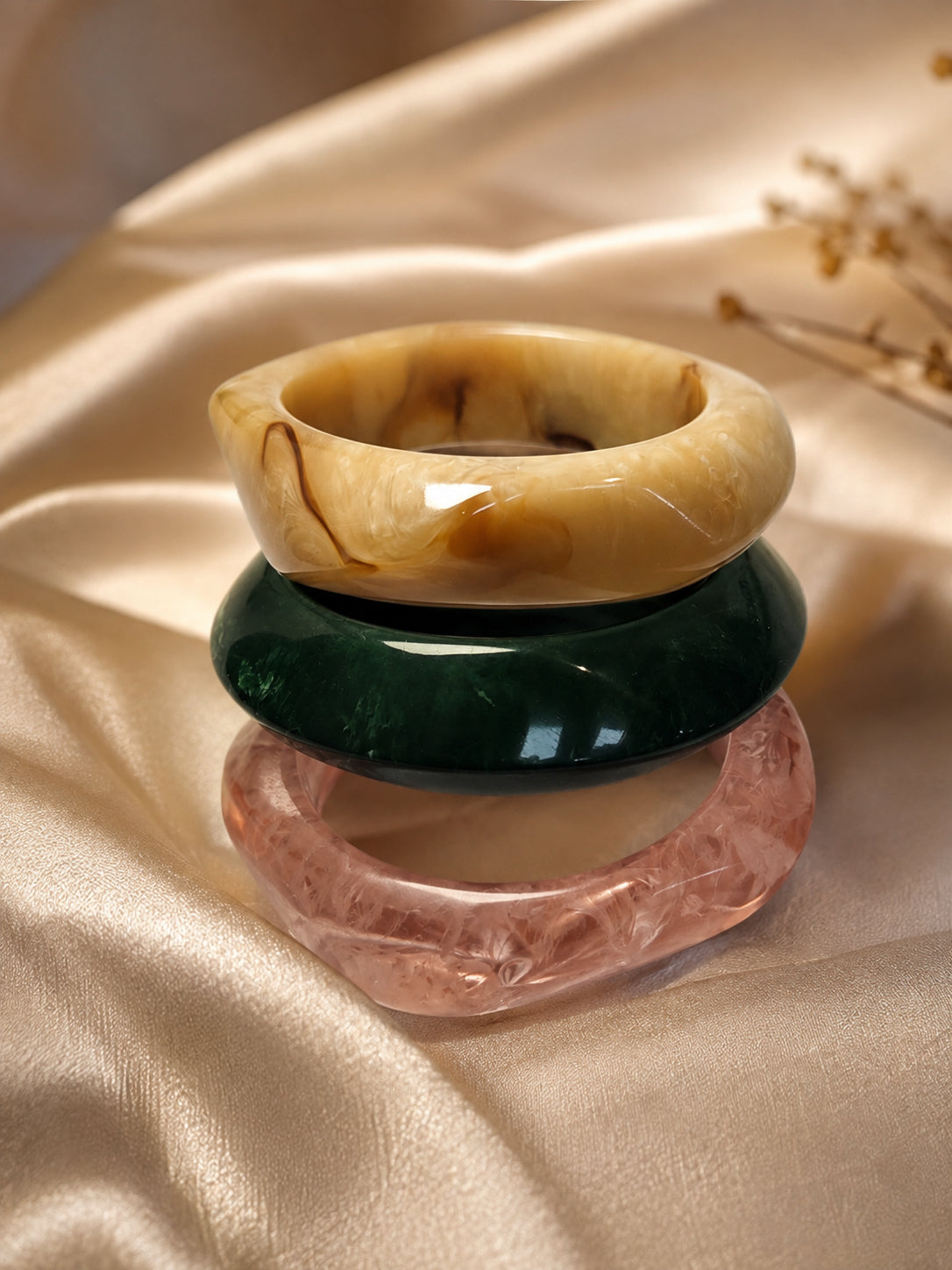 Amber, green, and rose marble resin bangles stacked on golden satin fabric with dried flowers